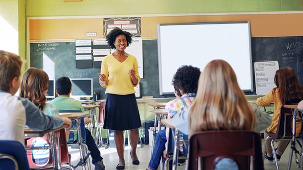 A woman stands in front of a classroom, engaging with students seated at desks.