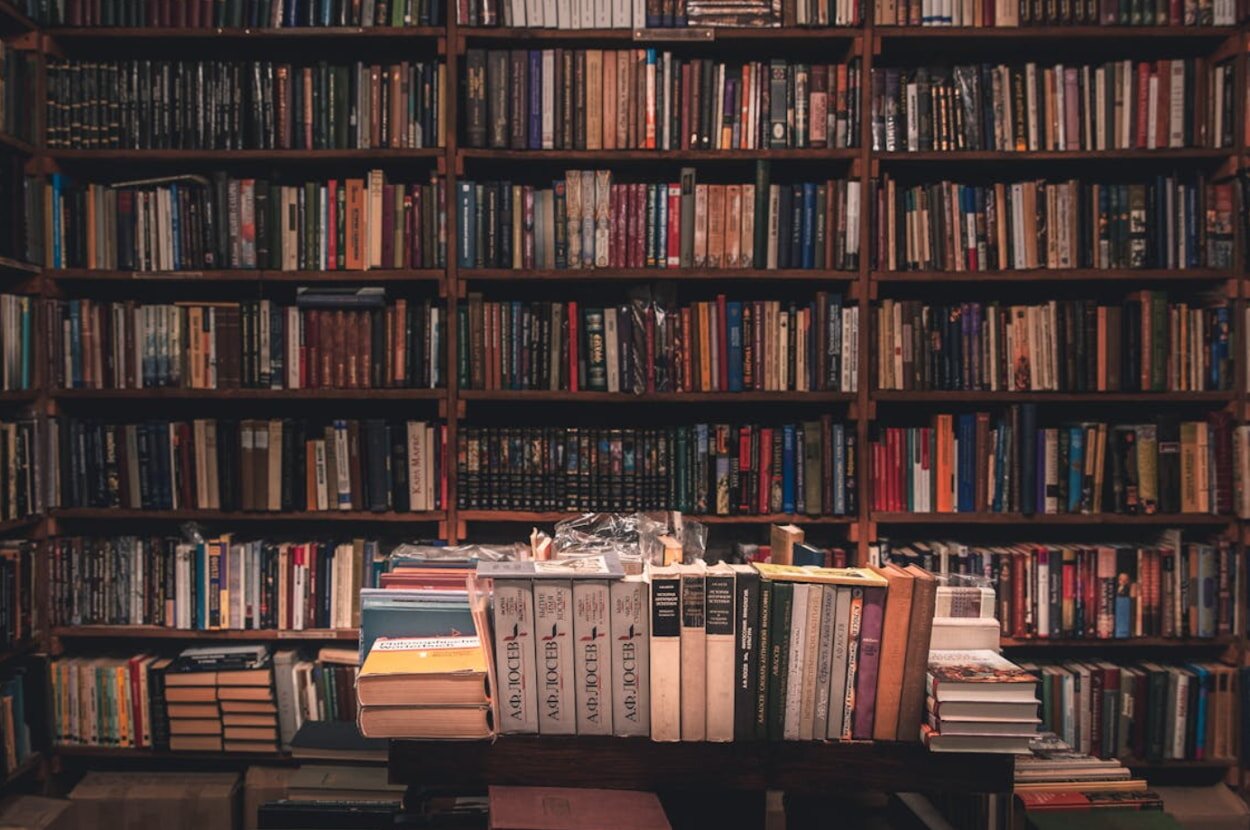 A table with several books on it, positioned in front of a tall bookshelf filled with more books.