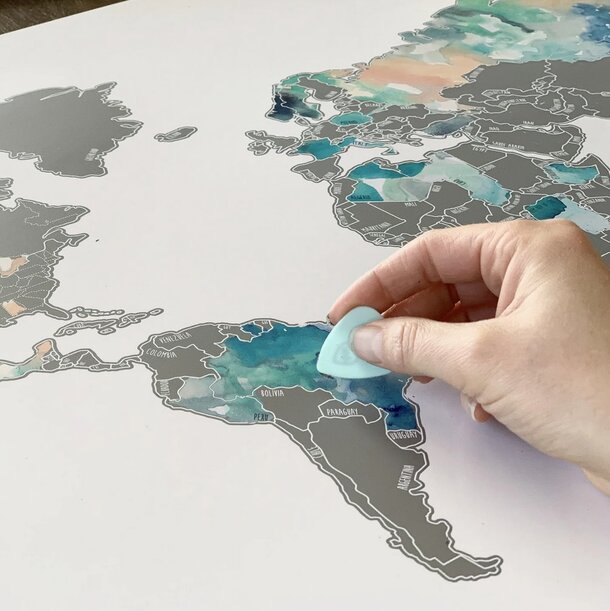 A hand scratches a silver map of the world, revealing colorful countries beneath. The map highlights Europe and South America, evoking discovery.