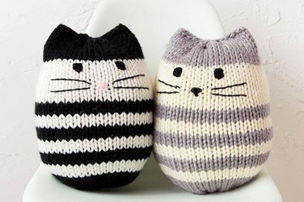 Two knitted cats are sitting side by side on a wooden chair, showcasing their colorful yarn patterns.