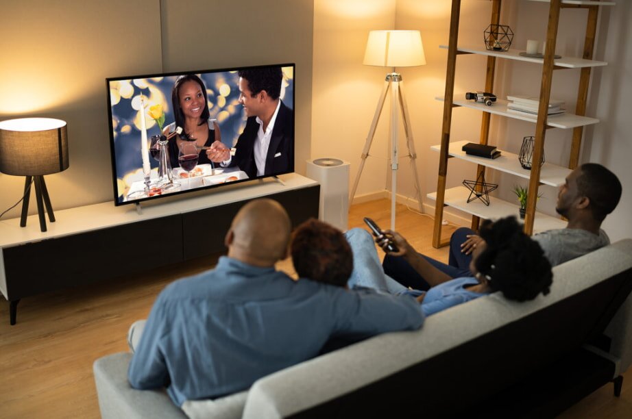 A family of four watches TV in a cozy, warmly-lit living room. The screen shows a couple dining. The mood is relaxed and intimate.