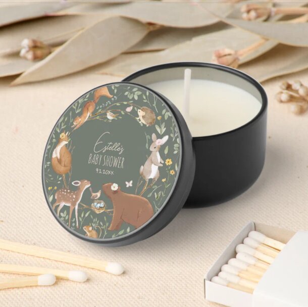 A black candle tin lid features woodland animals like a bear, deer, and rabbit surrounded by flowers, with the text "Estelle's Baby Shower." Matches lie nearby.
