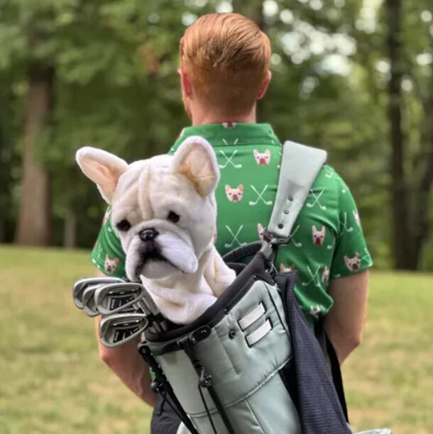 A man stands beside a golf bag, playfully holding a small dog that peeks out from inside the bag.