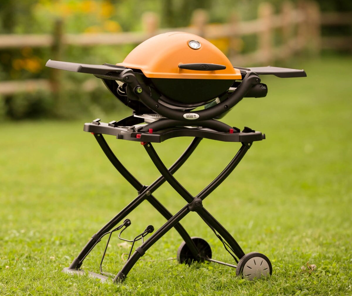 A compact orange grill on a foldable black stand sits on a lush green lawn. The background features a blurred wooden fence and yellow flowers.