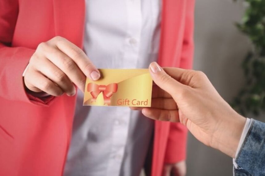 A woman presents a gift card to another woman, both smiling in a friendly exchange.