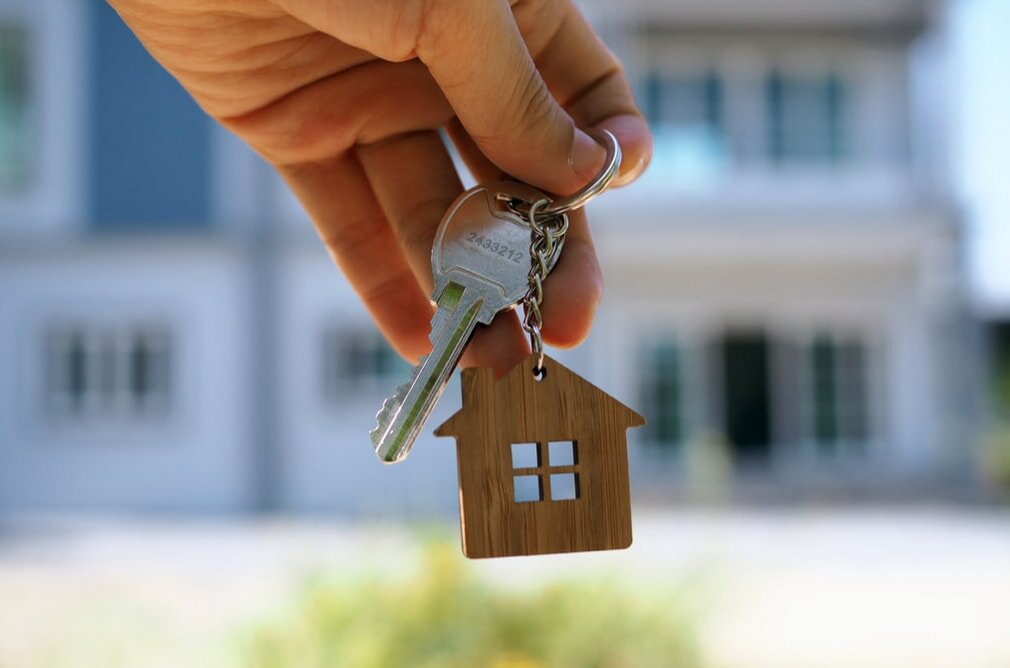 Hand holding a keychain with a house-shaped charm in front of a blurred home background, symbolizing new homeownership, evokes a hopeful mood.