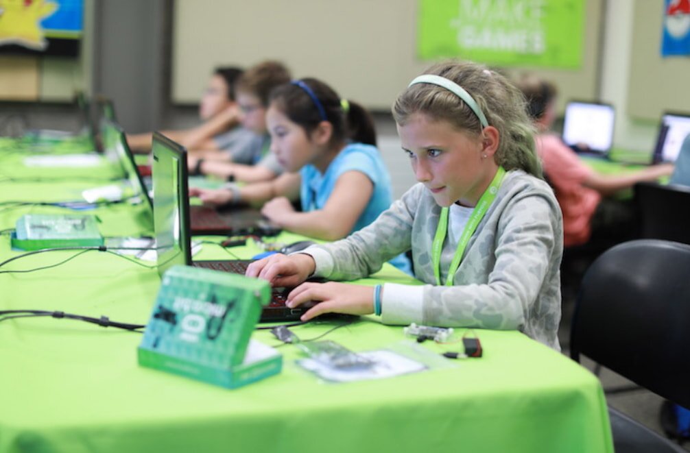 A girl sitting at a table, focused on her laptop, with a notebook and pen beside her.