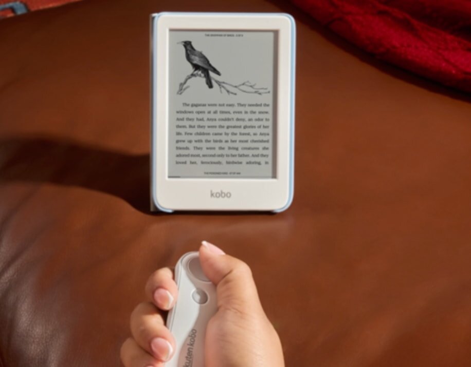 A hand holds a Kobo remote in front of an e-reader displaying a page with a bird illustration. The scene is cozy on a brown leather couch.