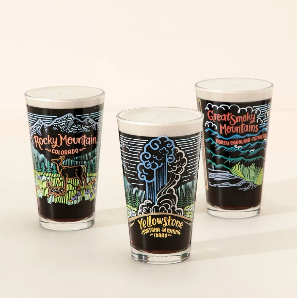Pint glasses printed with national park designs, each beside its gift box.