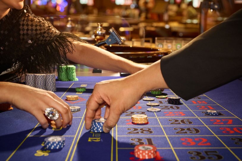 Hands reaching over a roulette table with colorful chips placed on numbers. One person wears a sparkling ring, suggesting a glamorous casino scene.
