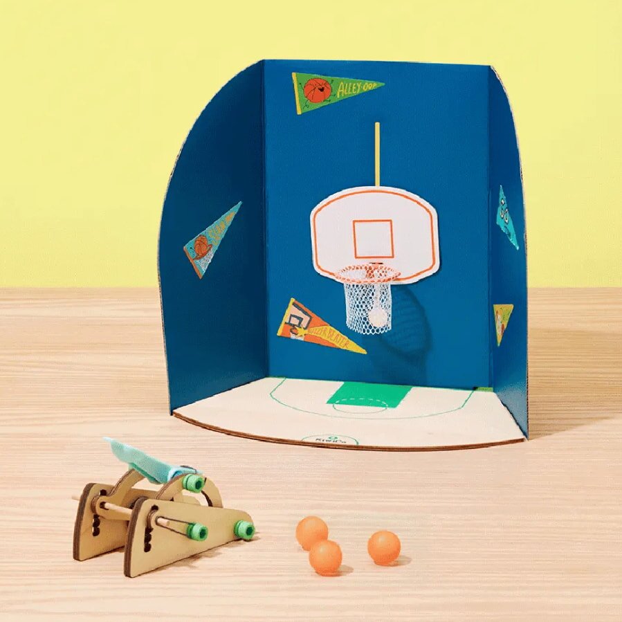 A mini basketball setup with a wooden catapult and orange balls on a light wood surface. A blue backdrop displays a hoop and colorful pennants.