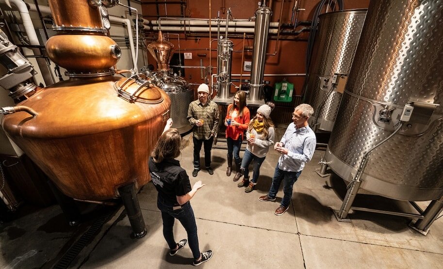 A tour guide explains the distillation process to four people in a distillery, surrounded by large copper and steel distillation tanks. The group appears engaged and curious.