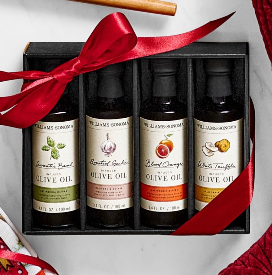 A gift set of four Williams-Sonoma infused olive oils, flavors: Aromatic Basil, Roasted Garlic, Blood Orange, White Truffle. Wrapped with a red ribbon.