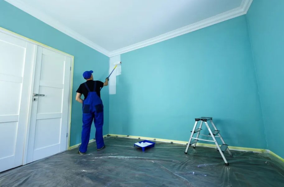 A person in blue overalls paints a light blue wall white with a roller. A step ladder and paint tray are on a plastic-covered floor in a tidy room.