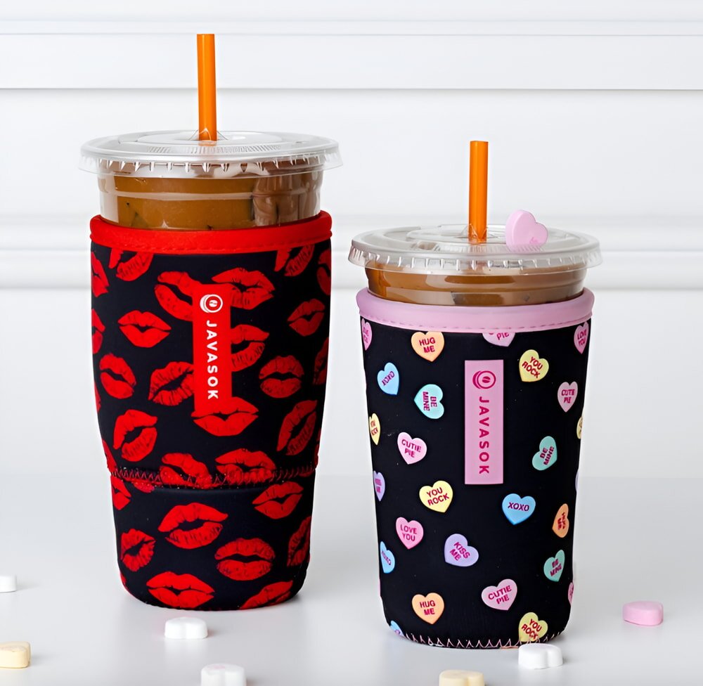Two iced coffee cups with orange straws are in colorful sleeves. One has red lips, and the other has candy heart designs, creating a playful, romantic vibe.