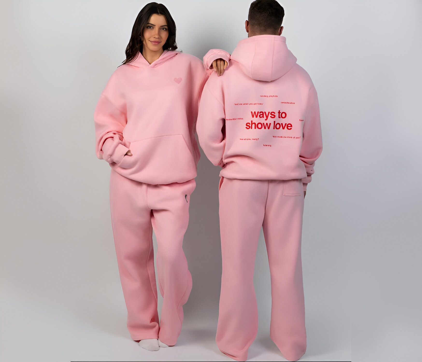 Two individuals wearing matching pink hoodies and sweatpants. The front has a simple heart, while the back reads "ways to show love" in bold red text. Mood is cozy.