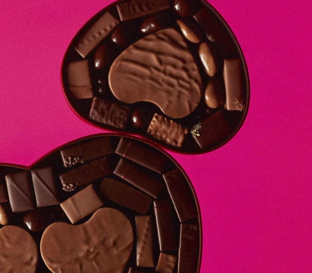 Heart-shaped chocolate boxes against a bright pink background. Boxes are filled with assorted chocolates, creating a sense of indulgence and luxury.
