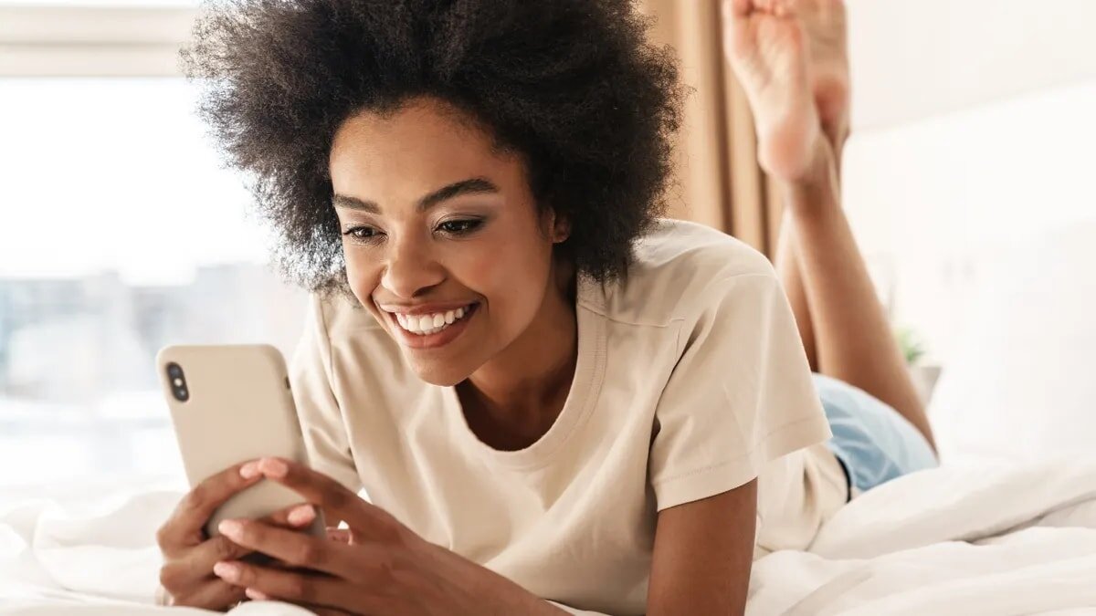 A woman happily lays on her bed, engaged with her phone, creating a warm and inviting atmosphere.