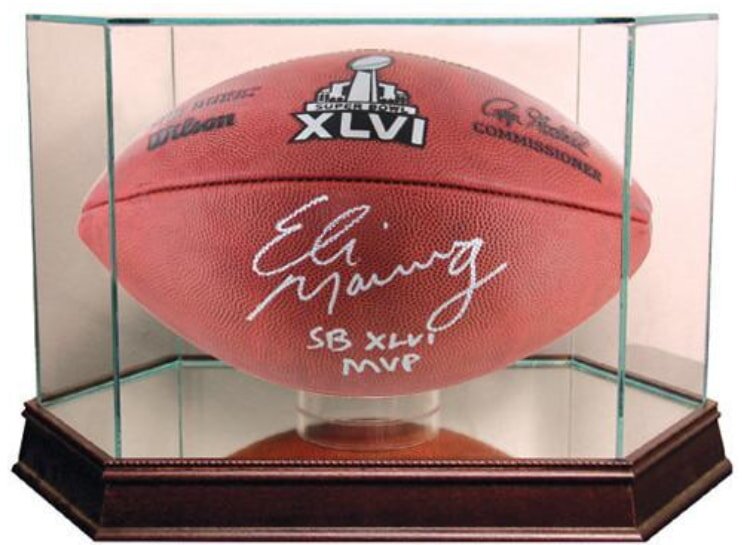 A signed football with "Eli Manning" and "SB XLVI MVP" displayed in a clear glass case with a wooden base. The ball features a Super Bowl XLVI logo.