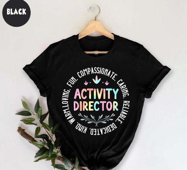 Black t-shirt on hanger with colorful "Activity Director" text, surrounded by words like "compassionate" and "fun," conveying positivity and dedication.