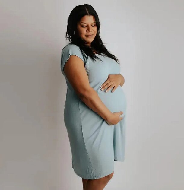 A pregnant woman wearing a blue dress stands gracefully, showcasing her baby bump with a serene expression.