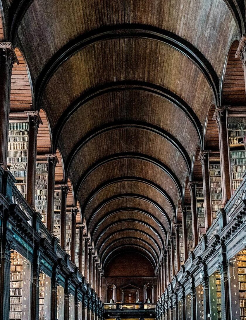Historic library hall with a high, arched wooden ceiling and towering bookshelves filled with books. The setting feels grand and serene.