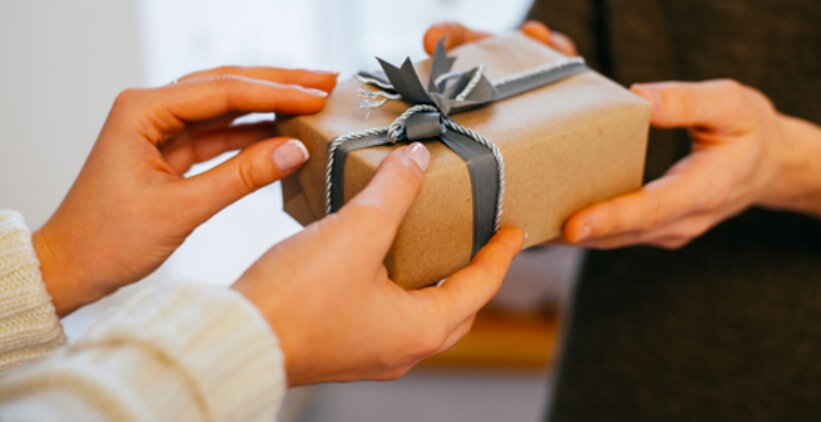 Two people exchanging a gift wrapped in brown paper with a gray ribbon. The hands and gift are in focus, conveying warmth and generosity.