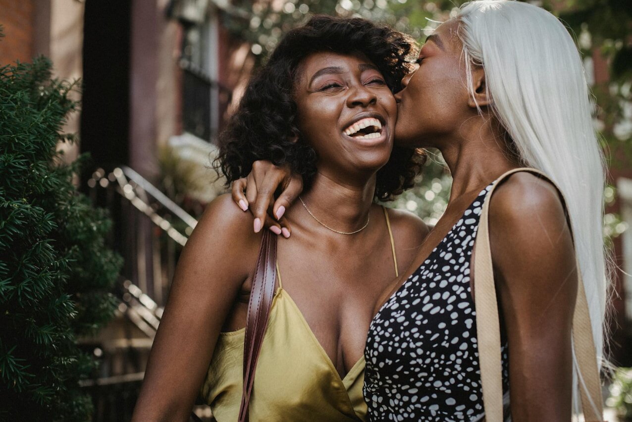 Two women share a kiss in a lively street, surrounded by urban scenery and passersby.