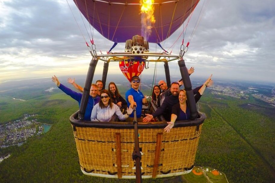 A group of smiling people enjoying a hot air balloon ride high above a sprawling green landscape and town, with a vibrant sky overhead.