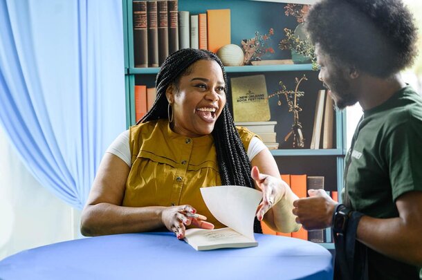 A woman and a man sit at a table, engaged in conversation while looking at an open book between them.