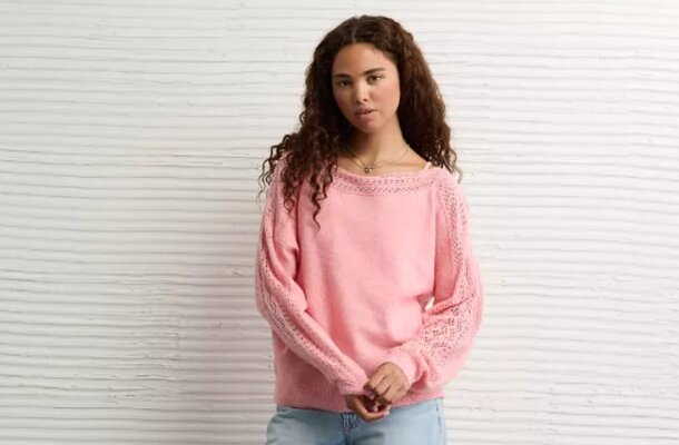 A woman wearing a pink sweater and jeans stands casually, smiling at the camera.