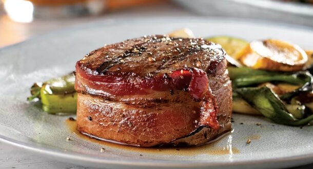 A plated steak topped with bacon, accompanied by a colorful assortment of vegetables.