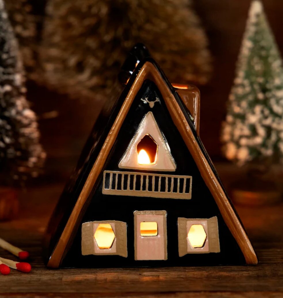 Cozy ceramic candle holder shaped like an A-frame cabin, glowing warmly with a lit candle inside. Snowy evergreen trees in the blurred background.