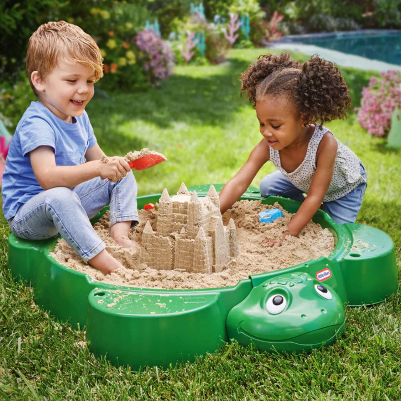 Two children happily playing in a sand pit, with a small frog nearby, enjoying a sunny day outdoors.