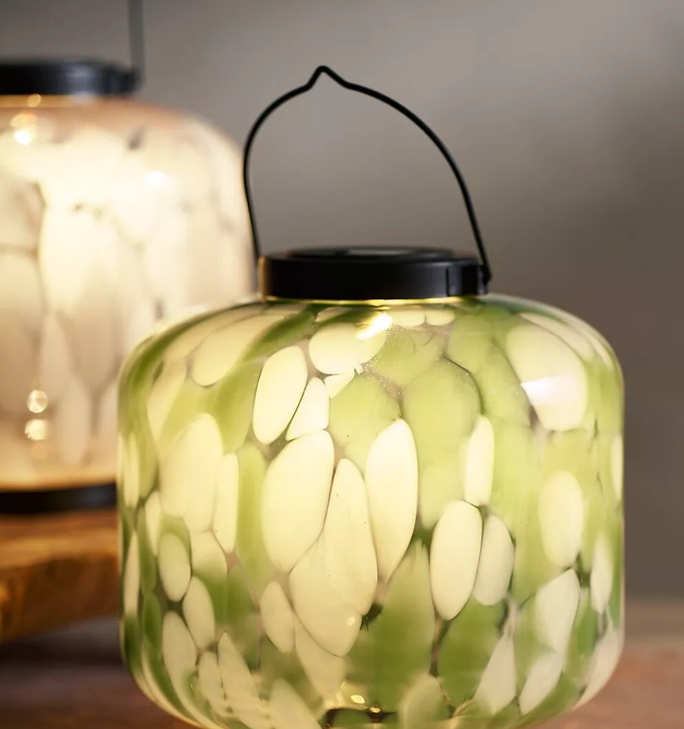 A decorative glass lantern featuring a green and white design, casting a soft glow in a dimly lit setting.
