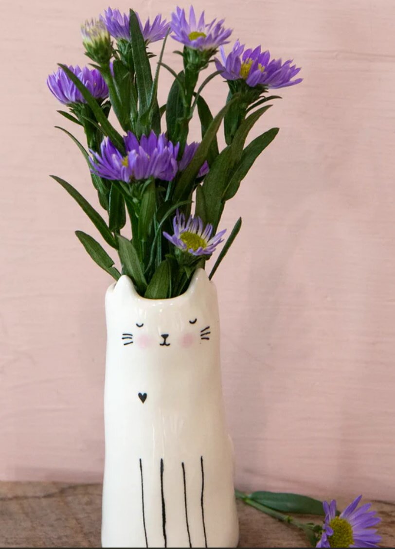 Ceramic vase shaped like a smiling cat with closed eyes, holding purple flowers and green leaves. The background is a soft pink wall.