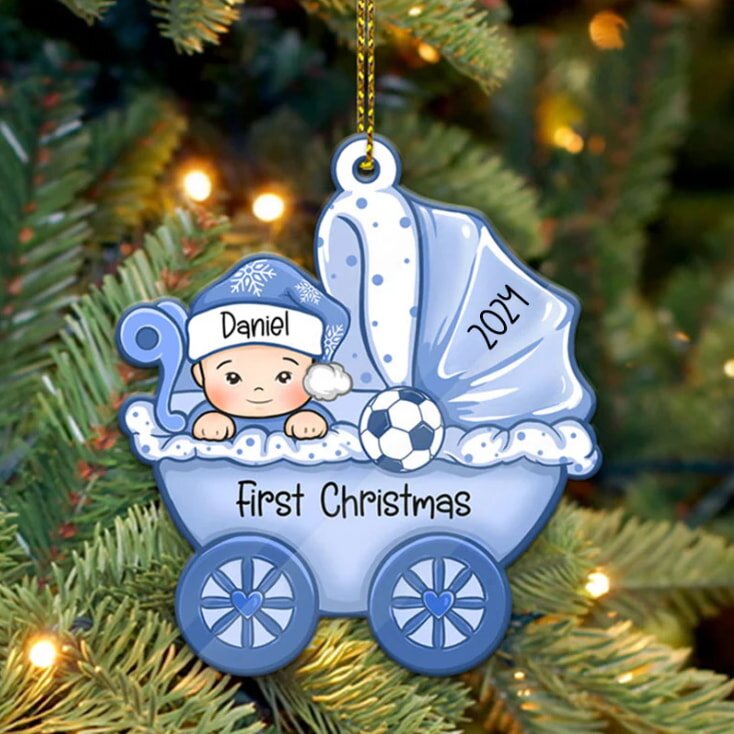Blue baby carriage ornament with a baby in a Santa hat, labeled "Daniel" and "2014." Text reads "First Christmas." Background: lit Christmas tree.
