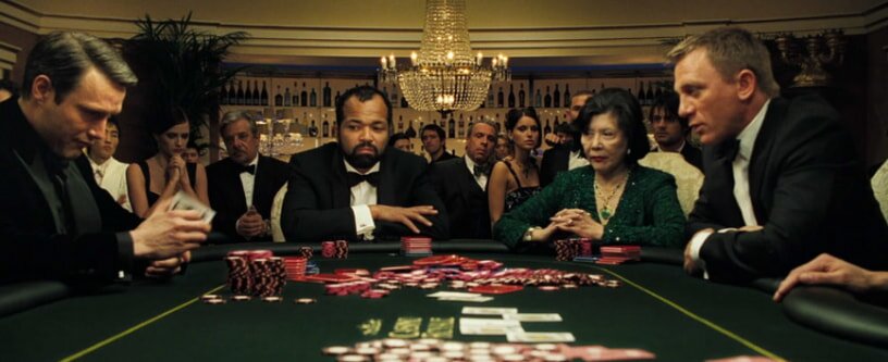 A tense poker game unfolds in a lavish casino setting. Four players sit at a green felt table with stacks of chips. An elegant chandelier hangs above, while an audience watches closely, creating a suspenseful atmosphere.