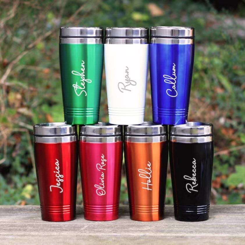 Seven personalized metal tumblers with names on a wooden surface outdoors