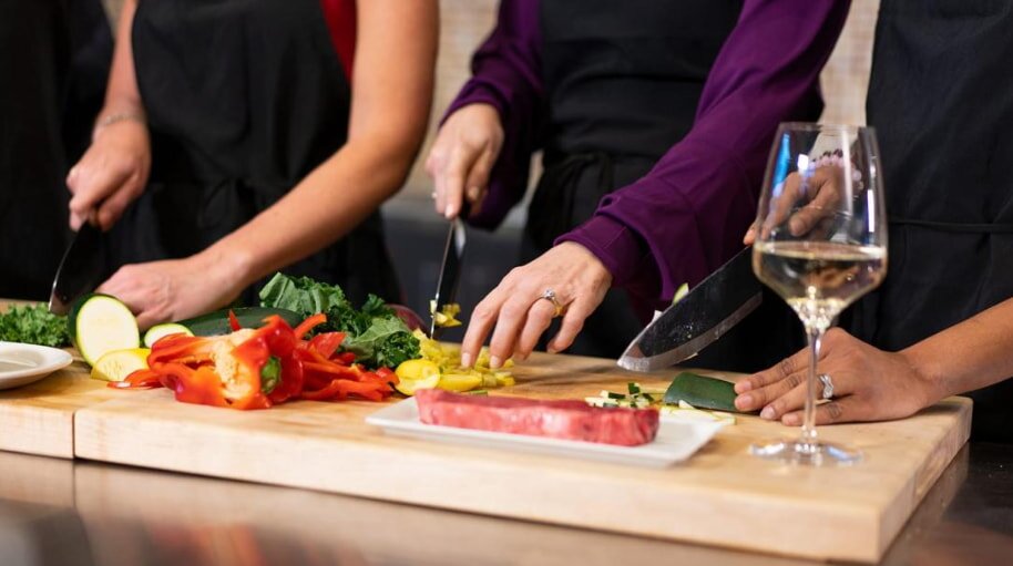 Three people in aprons slice vegetables on a wooden board. A glass of white wine is on the table, conveying a collaborative and relaxed cooking atmosphere.