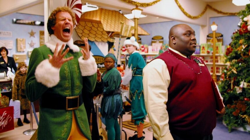 A man dressed as an elf stands next to a man dressed as a Christmas tree, both smiling in a festive setting.