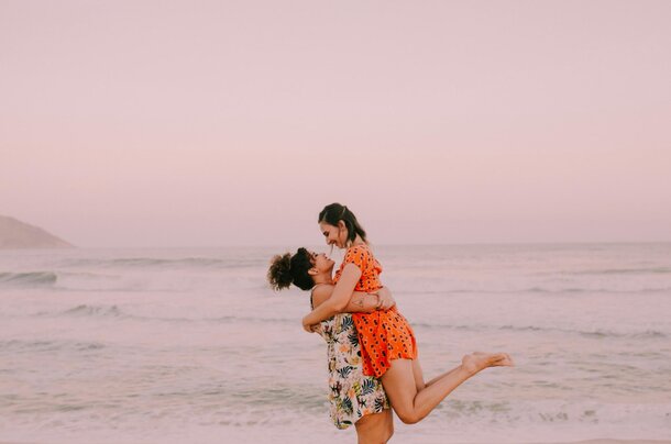 Two women embrace on a beach at sunset, with vibrant orange and pink hues illuminating the sky behind them.