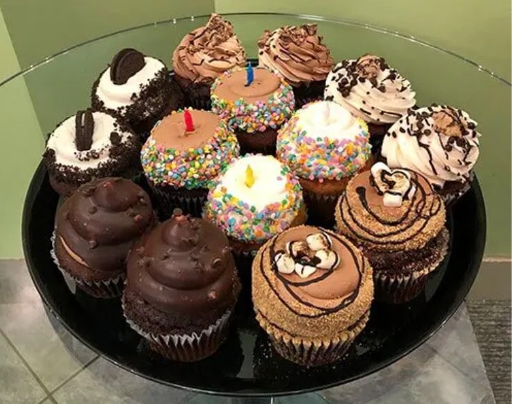 A platter of assorted cupcakes with colorful frosting and toppings such as sprinkles, chocolate, and cookies, arranged appealingly on a black tray.
