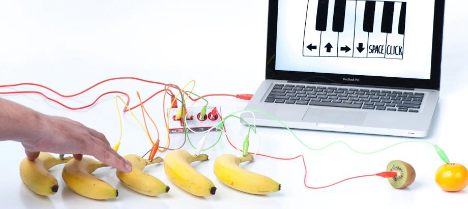 Hand touches bananas connected with wires to a laptop showing a piano keyboard on its screen. A kiwi and orange are also connected. Innovative, playful tech setup.