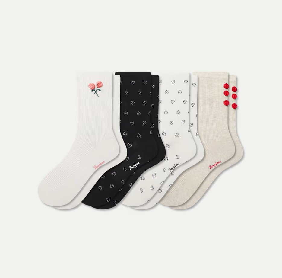 A set of five colorful socks featuring various designs: white with pink flowers, black with hearts, white with small hearts, beige with red buttons. Playful and cozy.