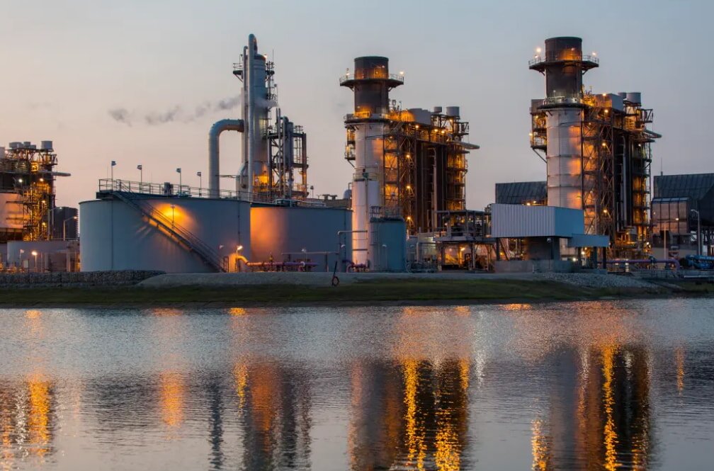 Industrial landscape at dusk, with illuminated gas refinery towers reflecting in a tranquil pond. The scene conveys a sense of technological sophistication.