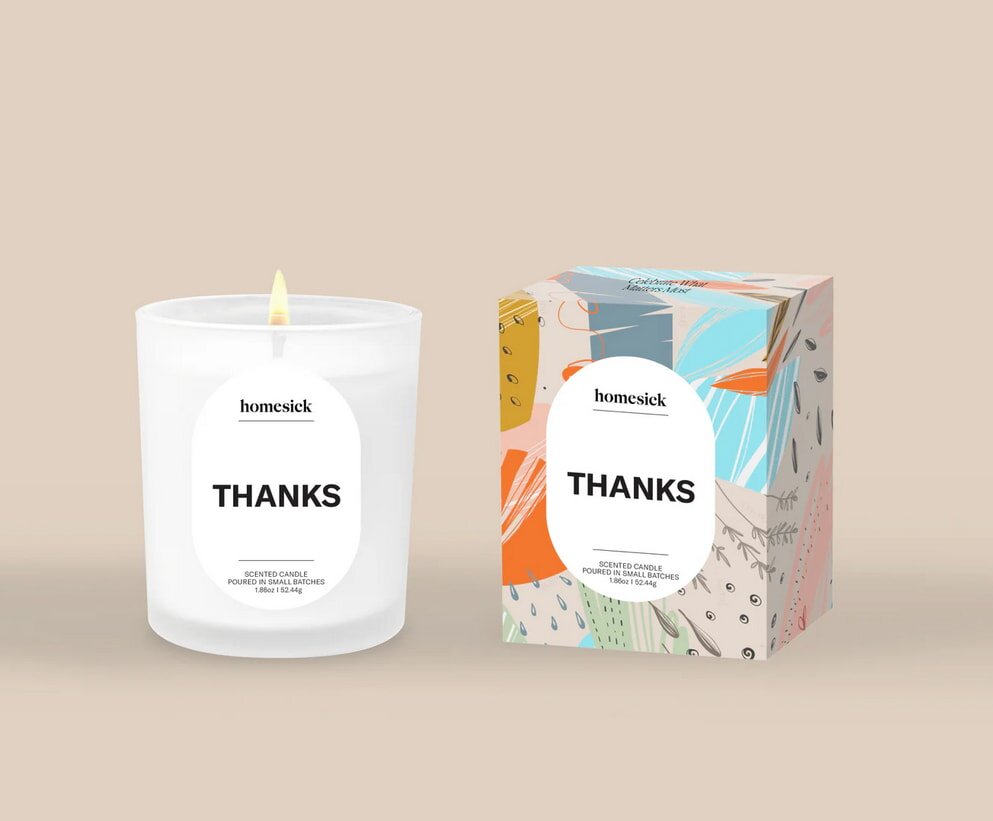 A lit white candle labeled "Homesick Thanks" stands next to a colorful box with abstract patterns and the same label, on a beige background. Calm and appreciative.