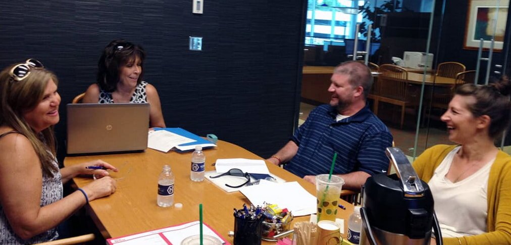 A woman in a black blazer hands keys to a man in a light shirt holding documents. They stand against a beige brick wall, conveying a professional exchange.