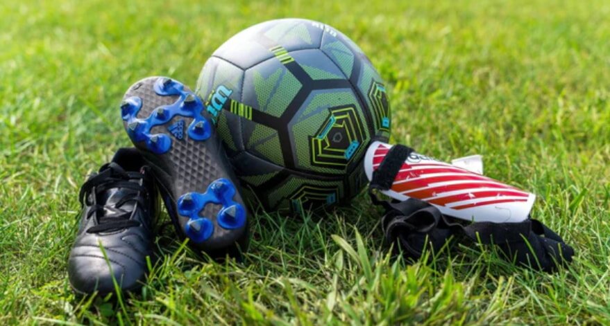 A soccer ball, black cleats with blue studs, red shin guards, and black gloves rest on a vibrant green grass field, evoking a sense of readiness.