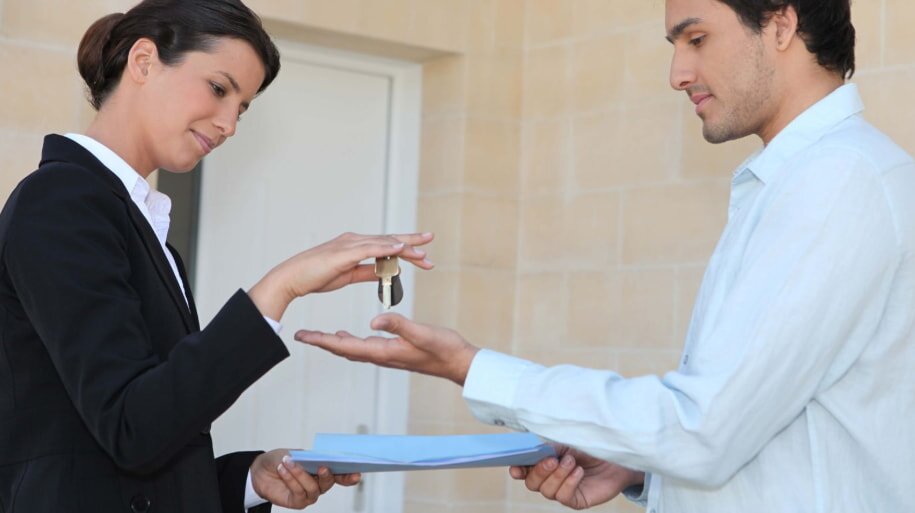 A woman in a black blazer hands keys to a man in a light shirt holding documents. They stand against a beige brick wall, conveying a professional exchange.